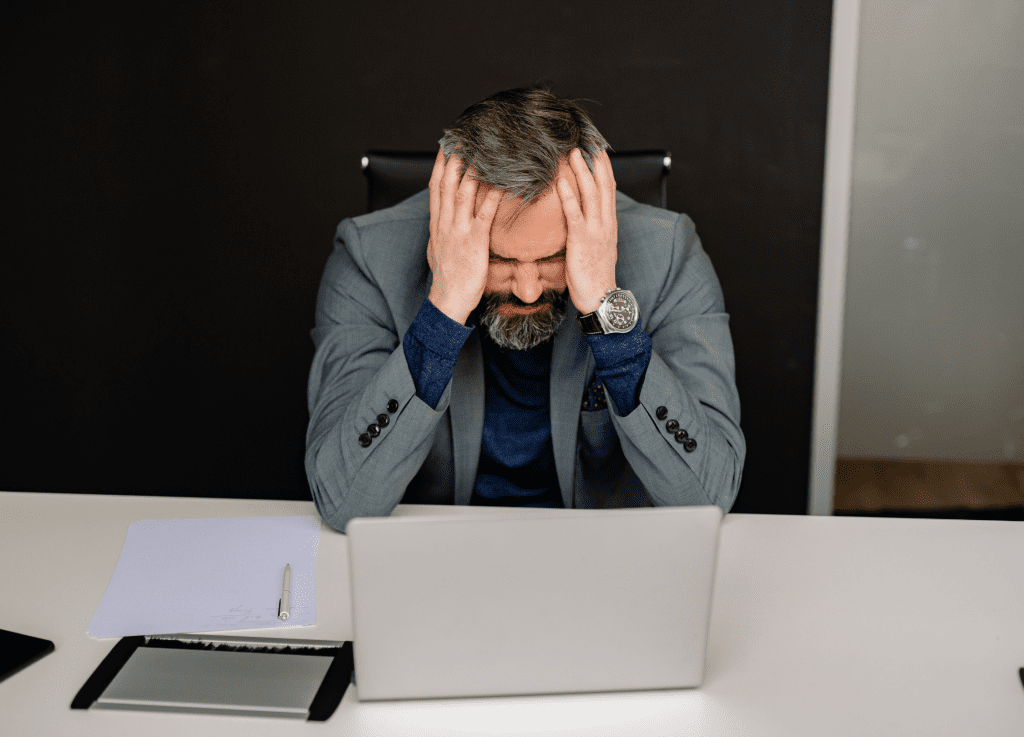 Overwhelmed American business owner sitting on desk surrounded by paperwork and laptop, stressed from work overload