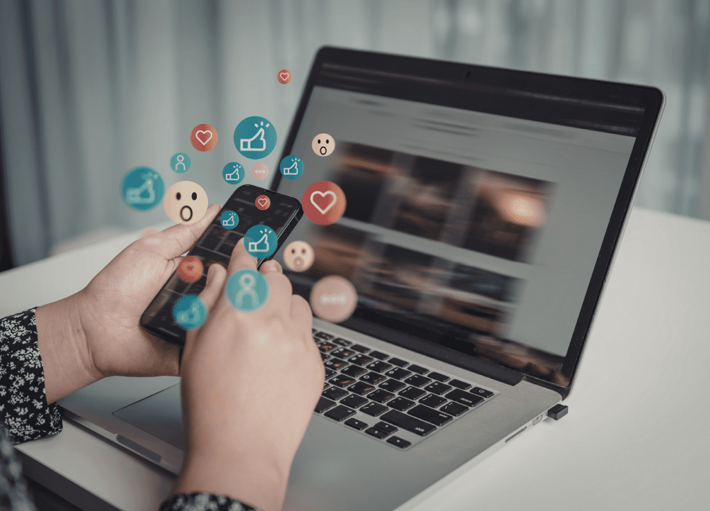Person using a smartphone over a laptop with floating social media icons like hearts, likes, and emojis representing digital engagement