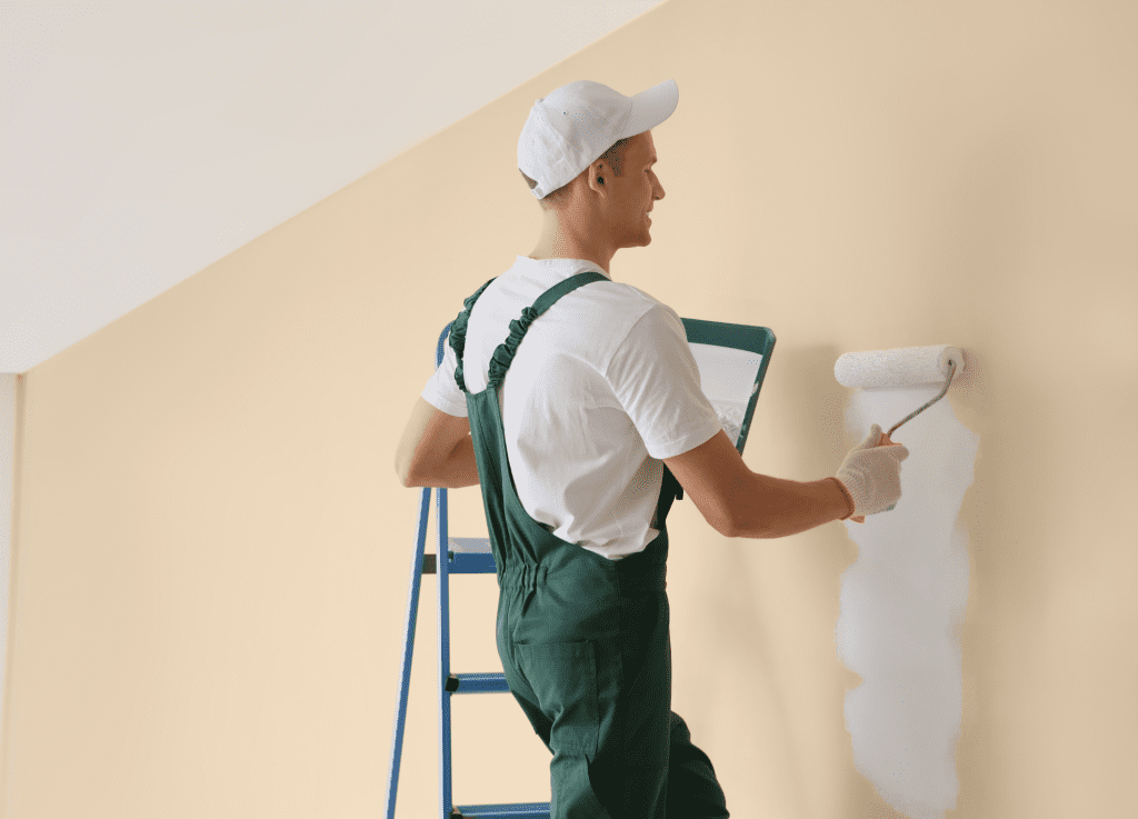 professional painter in green overalls and a white cap using a paint roller on a beige interior wall while standing on a ladder