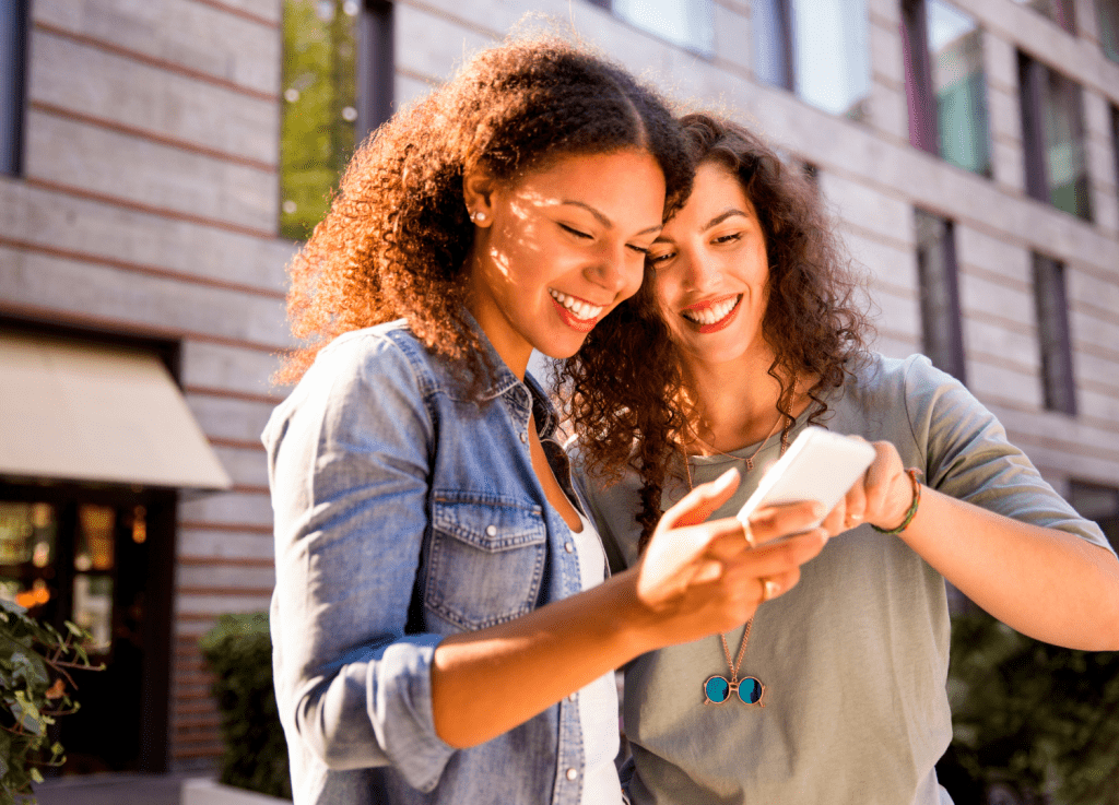 Two smiling young women standing outdoors and looking at a smartphone together