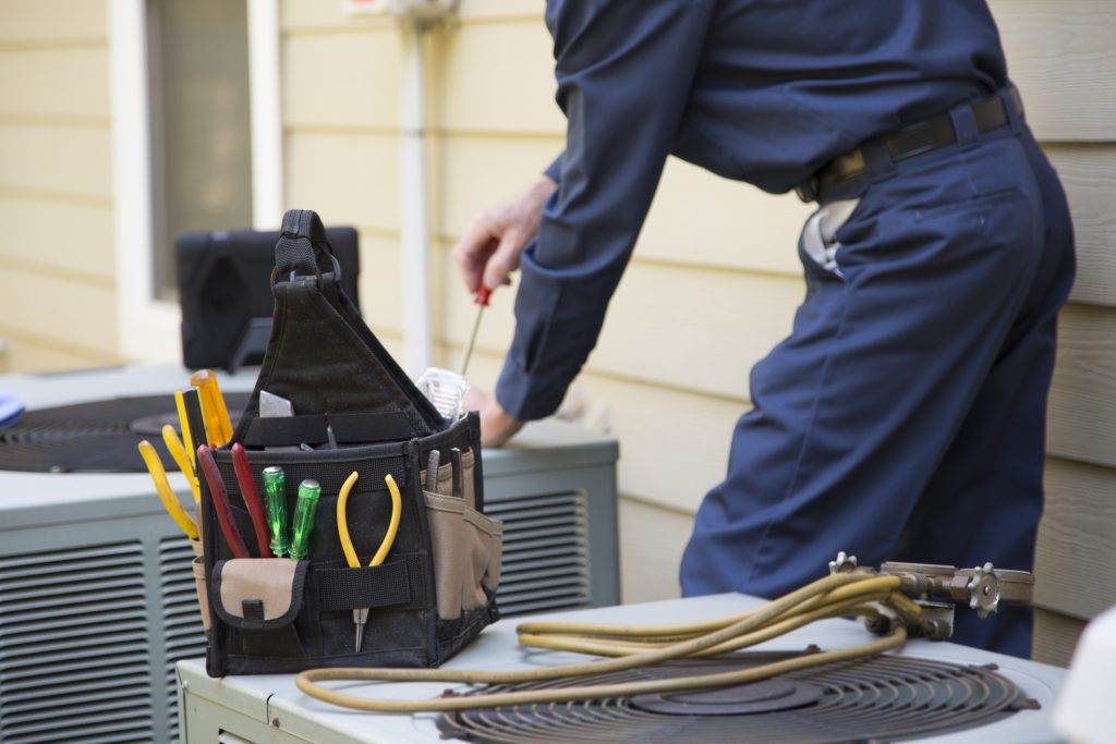 HVAC technician in a blue uniform repairs an outdoor air conditioning unit, with a black tool bag filled with screwdrivers and pliers in the foreground