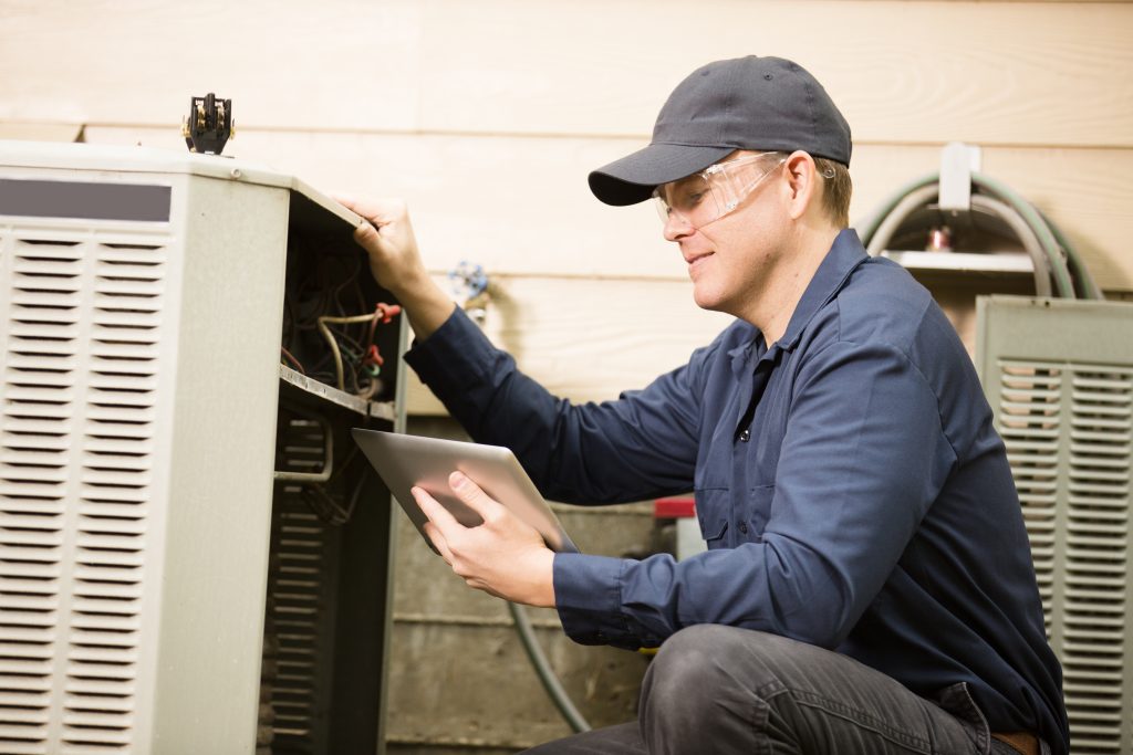 HVAC technician wearing safety glasses and a cap uses a digital tablet while inspecting the internal components of an outdoor air conditioning unit