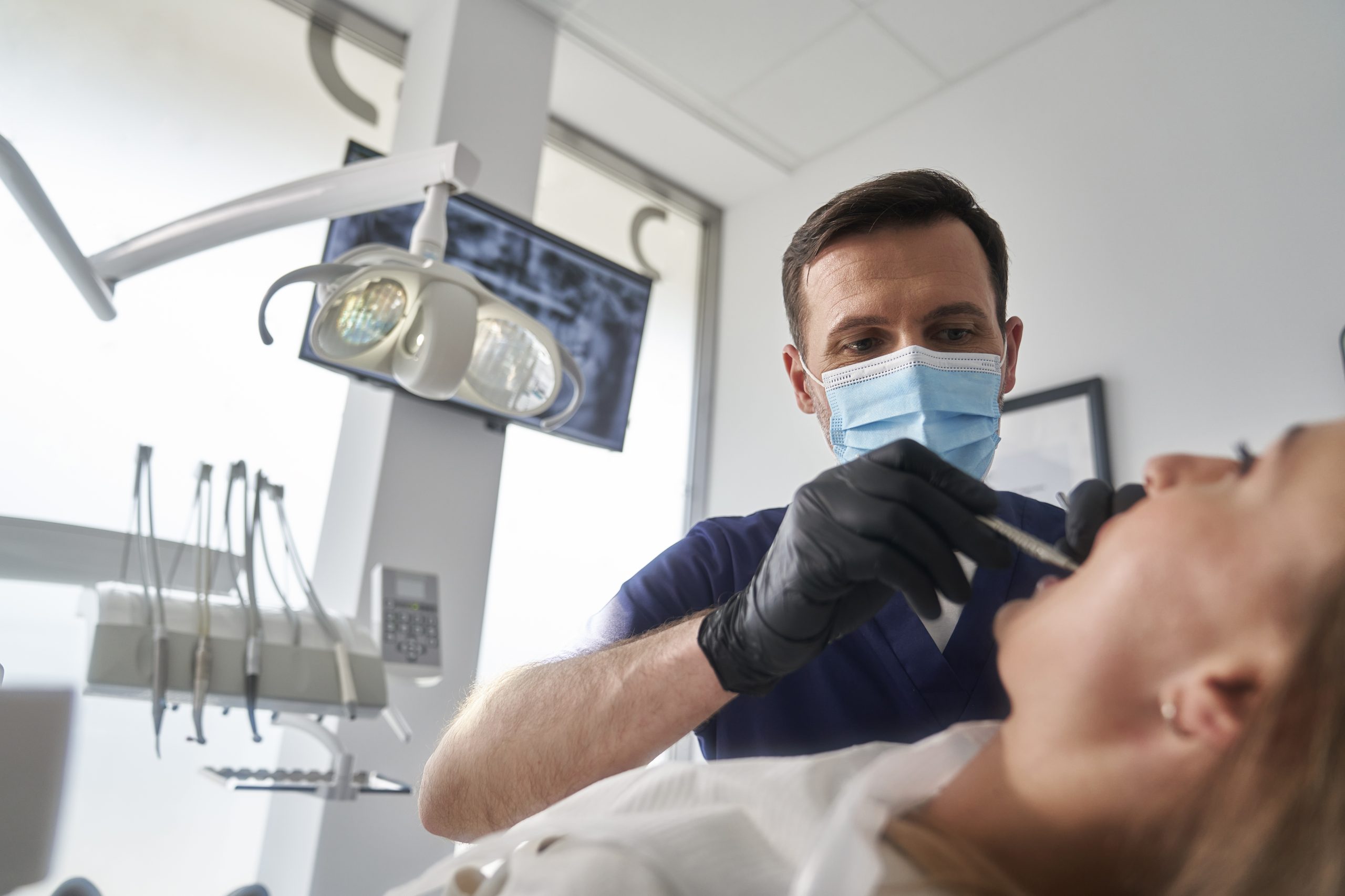 A professional male dentist wearing a face mask performs an oral exam on a patient in a modern dental office setup