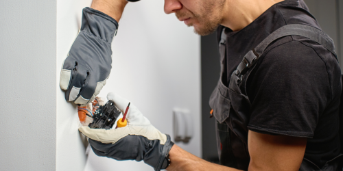 electrician wearing work gloves and overalls installing a new electrical outlet in a white wall
